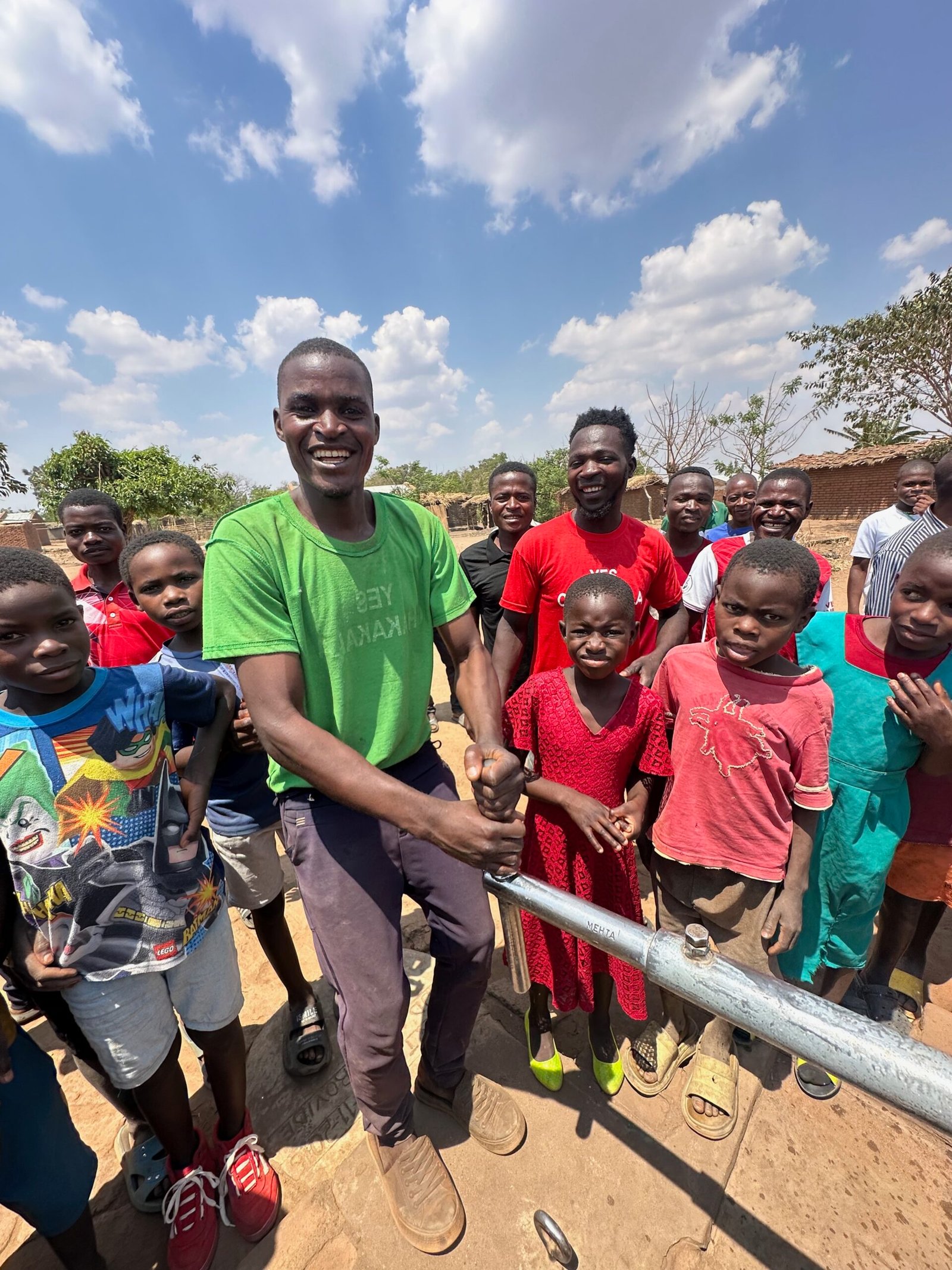 Smiling man in a green shirt shakes hands with someone at a metal barrier, surrounded by children outdoors on a sunny day with a blue sky.