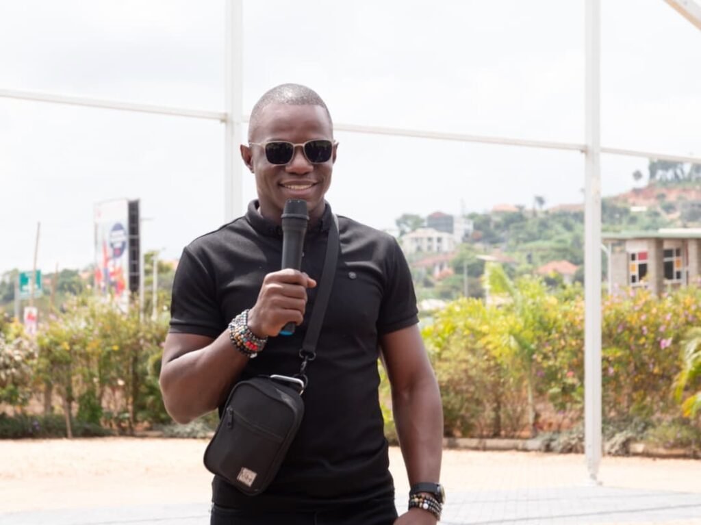 A man in a black polo holding a microphone, wearing sunglasses and a small black crossbody bag outdoors at an event.