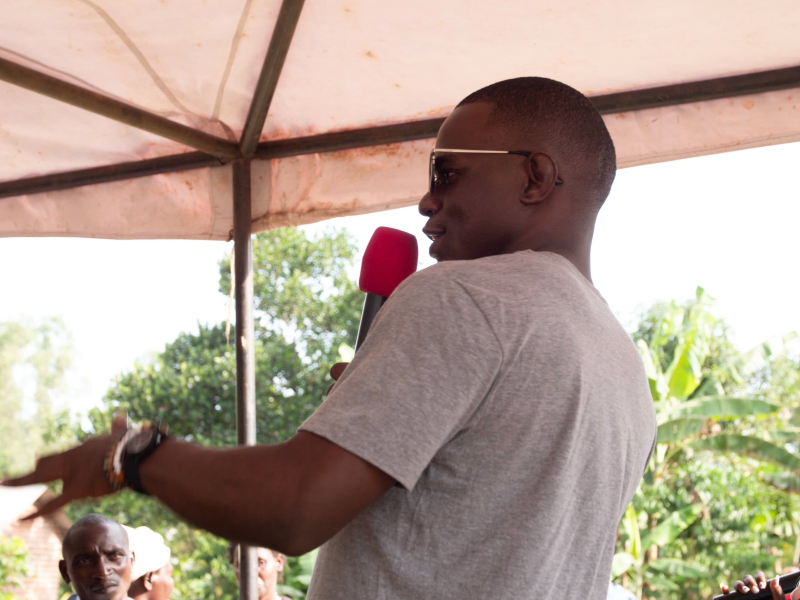 Man wearing sunglasses speaks into a red microphone under a canopy at an outdoor event.