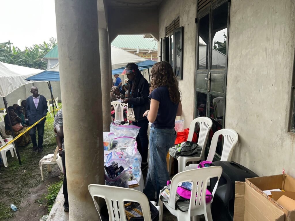 People gather at an outdoor aid event under a concrete walkway, with plastic chairs, boxes, and scattered supplies visible.