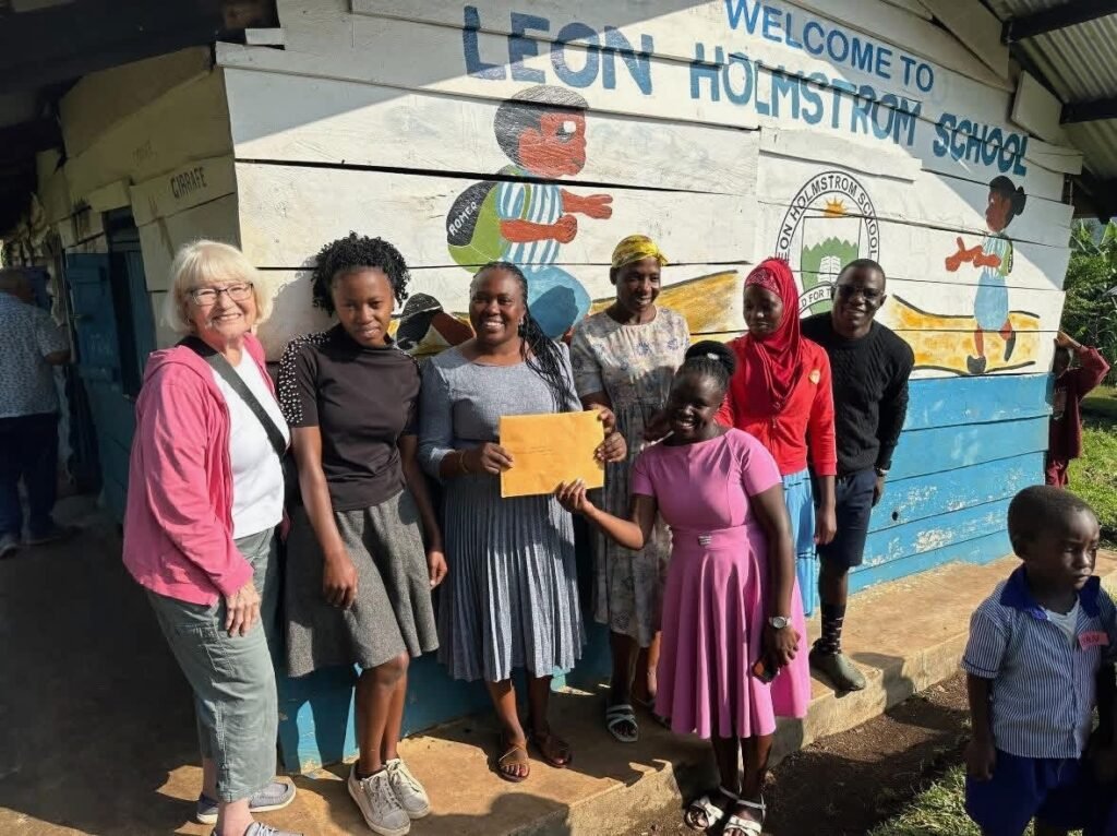 Group of adults and children pose outside a muraled school building, as a girl in pink hands an envelope to a woman in a gray dress.