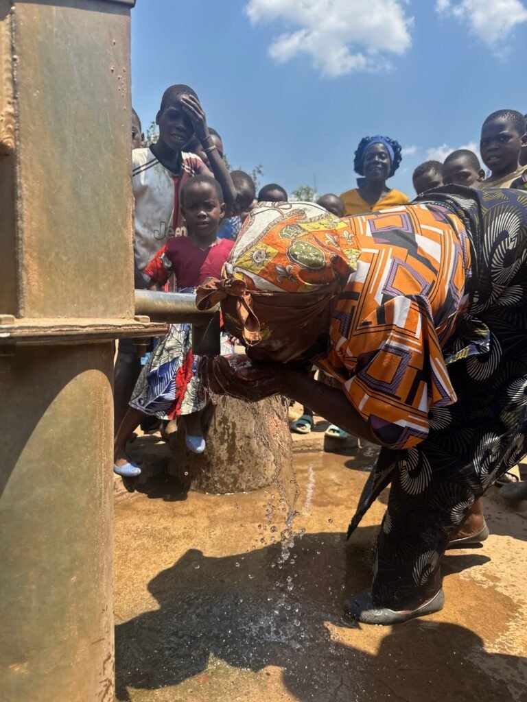 People gather around a large outdoor water pump as a woman in a bright patterned dress leans over to wash or collect water, while children stand nearby and watch under a clear blue sky.